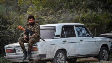 A volunteer fighter sits on a car as cannons fire nearby