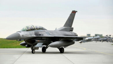 A US Air Force F-16 fighter jet is seen grounded on a runway. Its identification, SP 481, is painted on its tail. A group of jets can be seen in the background, too out of focus to identify which kinds.