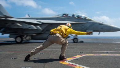 A military personnel is seen guiding a jet plane in what appears to be an on-sea landing system. The personnel is wearing a yellow jacket and brown khakis. The fighter plane is colored gray. The background is a cloudy blue sky, with the ocean water below.