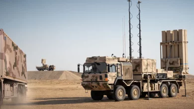 An IRIS-T SLM air defense system is seen in what appears to be a dry/desert landscape. Some sand can be seen being blown by the wind. The self-launching system itself is seen propped up and mounted on the back of an 8x8 military truck. The truck is painted in camouflage that matches the dry brown color of the environment. The background is a clear but dull blue sky.