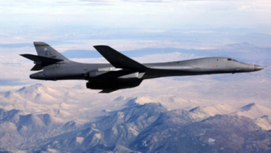 A black B-1B Lancer bomber is seen flying above a mountainous region. The mountains' land have colors ranging from forest's greens to desert yellows and whites. The sky above is cloudy and white.
