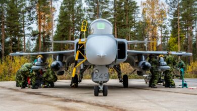 A Gripen C-series plane is seen being serviced by uniformed personnel while parked in the ground. All seven personnel are wearing green camouflage uniforms. The plane is painted gray with the number "249" painted in darker gray right below the front of the cockpit. The background is a forested area with trees and shrubbery.
