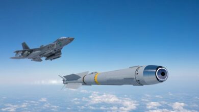 An artist's rendition of a KF-21 Boramae fighter jet flying above the clouds and firing off an IRIS-T missile. The jet plane, seen on the far left side of the image, is painted gray. The missile has a similar color scheme, seen jutting out white flames from its back as it propels itself forward. The background is a blue sky with white light clouds seen below.