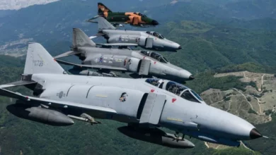 Four F-4E Phantom jet planes are seen flying over a green landscape in South Korea. Three of the planes are painted gray, while the one in the far left is painted camouflage green and orange.