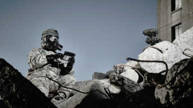 A soldier wearing camouflage fatigues and a General Service Respirator mask is seen using a wired device the size of a small tablet in a pile of rubble.