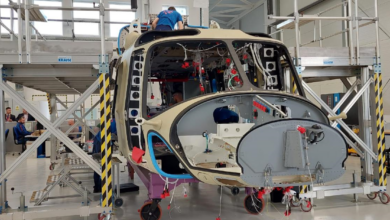 An AW149 Helicopter is seen in the middle of production. It has incomplete parts, but the shape of the front of the aircraft already resembles that of a usual helicopter. Wires and exposed framework are seen on the vehicle. A worker wearing a blue shirt is seen coming up out of its roof. Other personnel are seen in the far back in what appears to be an office space with desks and cubicles.