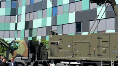 A Thales ControlMaster 200 is seen displayed in front of a building with window panes in gray, light green, and dark green colors. The system's truck is painted in deep green and brown camouflage colors. Ukrainian president Volodymyr Zelenskyy is seen walking on the bottom left side of the image with two other people wearing suits.