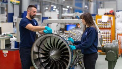 Two individuals wearing navy blue shirts with the Rolls-Royce logo on the left side of the chest are see inspecting a Typhoon aircraft engine. They appear to be inside an industrial building, with steel equipment and finishing in the blurred background.