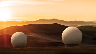 Two of Northrop Grumman’s relay ground stations are seen over the horizon in a field. The image has an orange tinge from the sun seen in the upper left side of the image. The white buildings are shaped like orbs.
