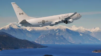 A Royal Canadian Air Force P-8A Poseidon aircraft is seen flynig over a large body of deep-blue water. In the far background, a snow-capped mountain range can be seen.