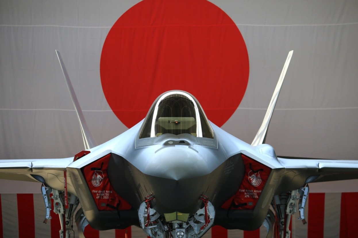 A silver-gray F-35 Lightning II jet fighter is seen taxied in front of an unfurled flag of Japan. The photo was taken front and center, displaying the plane's front profile.