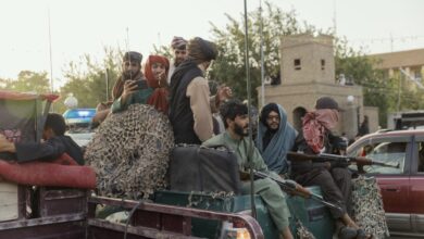 Taliban personnel holding guns and phones while riding on the back of a truck