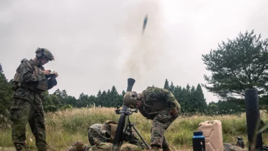 Soldiers conduct a live mortars firing during a field exercise, with one round captured mid-launch from the tube as the crew coordinates targeting and safety procedures in a grassy training area