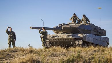 Soldiers stand and communicate beside a Leopard battle tank positioned on open terrain during daylight, with crew members visible on the turret and the main gun facing forward under a clear sky