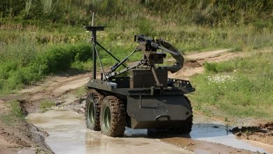 Armed ground robot driving through a muddy dirt road during a field test, equipped with a mounted weapon system and sensors while navigating shallow water in a rural environment
