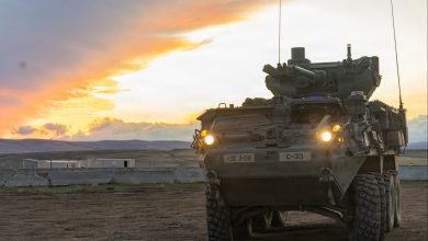 Stryker armored combat vehicle positioned on a dirt training range at sunset, headlights illuminated, with its remote weapon station visible against an open landscape.