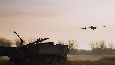 Rheinmetall’s LUNA NG unmanned aerial vehicle flying during a Bundeswehr field trial, operating alongside a ground-based air defense system in a rural training area at dusk