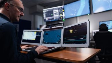 Thales technician operating a tri-screen laptop workstation displaying maritime surveillance, tracking, and tactical data feeds in a dimly lit defense operations center