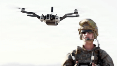 U.S. Army soldier in camouflage and tactical helmet alongside an airborne quadcopter drone equipped with a camera payload, supporting a story on converting vehicle periscope ports into drone launch ports