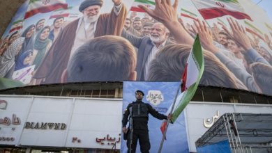 An Iranian police officer holds a national flag on a stage alongside a Persian script reading 'Flag bearer', under a giant billboard depicting an AI-generated portrait of Iran's late Supreme Leader Ayatollah Ali Khamenei in downtown Tehran, Iran, on April 23, 2026.