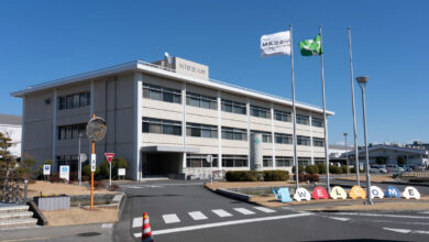 Nissan Motor's Oppama plant in daylight, building situated beside road with flag bearings in front.